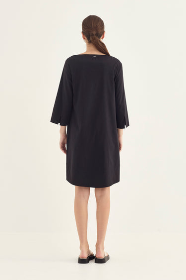 A woman viewed from the back wearing a loose black dress by Verge with three-quarter sleeves and black flat shoes, standing against a plain light background.