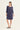 A woman modeling a loose, knee-length dress in a muted periwinkle or dusty purple shade. The dress by Verge has three-quarter sleeves and a relaxed silhouette, paired with black slide sandals, set against a plain off-white studio background.