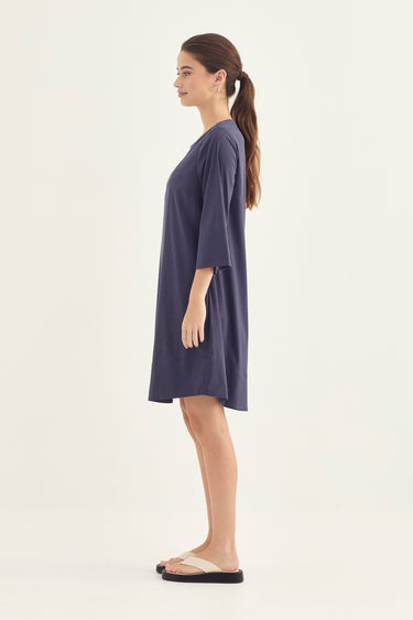 A woman in profile wearing a loose, short-sleeve navy blue tunic dress by Verge and beige slide sandals, standing against a plain off-white studio background.