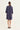 A woman viewed from the back wearing a loose, knee-length navy dress by Verge with three-quarter sleeves and black flat shoes, standing against a plain off-white studio background.