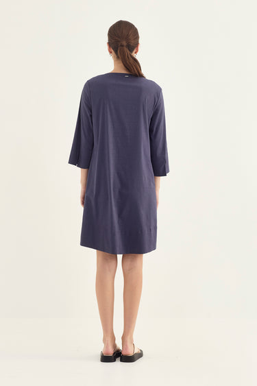 A woman viewed from the back wearing a loose, knee-length navy dress by Verge with three-quarter sleeves and black flat shoes, standing against a plain off-white studio background.