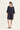 A woman modeling a loose, short-sleeve navy blue dress by Verge with a wide neckline and straight silhouette, paired with black slide sandals, against a plain off-white studio background.