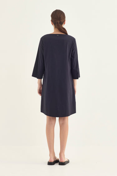 A woman viewed from the back wearing a dark navy blue shift dress by Verge with three-quarter sleeves and black flat shoes, set against a plain off-white studio background.
