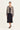 A model stands against a plain light background wearing a black midi skirt, a black top, and a cropped long-sleeve jacket by Verge with a beige and black snakeskin pattern.