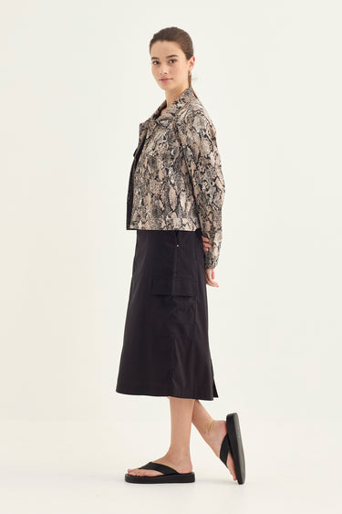 A model is shown in profile wearing a cropped, long-sleeve jacket with a beige, black, and gray snake print, paired with a black midi skirt by Verge and black flip-flop sandals, against a plain light background.