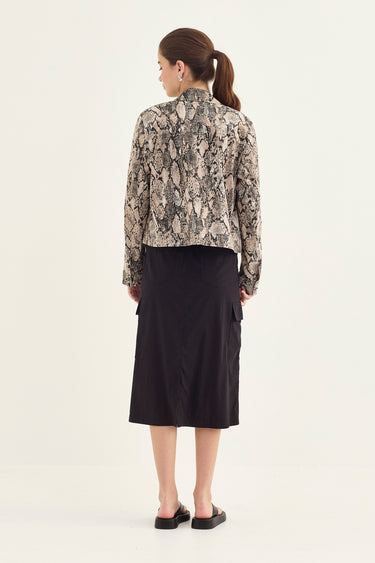 A woman shown from the back wearing a cropped long-sleeve blouse with a beige and gray snakeskin pattern and a black midi skirt by Verge, against a plain off-white studio background.