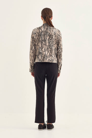 A woman viewed from the back wearing a cropped long-sleeve shirt with a gray and beige snakeskin pattern, paired with black trousers by Verge and black sneakers, against a plain white studio background.