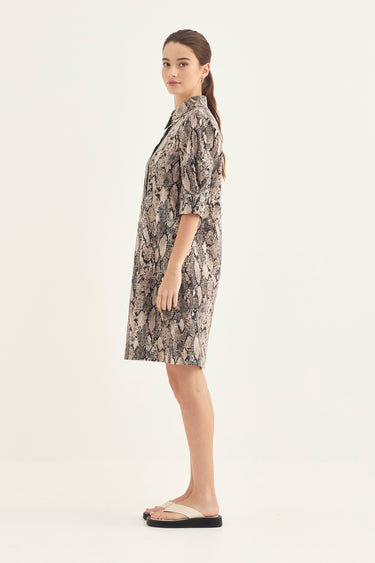A woman stands in profile wearing a short-sleeve collared shirt dress by Verge with an abstract black, gray, and beige print, paired with beige slide sandals, against a plain off-white studio background.