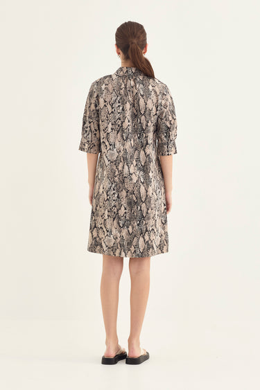 A woman viewed from the back wearing a short-sleeve shirt dress by Verge with a beige, gray, and black snakeskin pattern, paired with black flat shoes, against a plain off-white background.