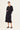 A model stands against a plain light background wearing a black long-sleeve collared jacket over a black top with a gold-toned embroidered chest design, paired with a black midi skirt by Verge and black slide sandals.