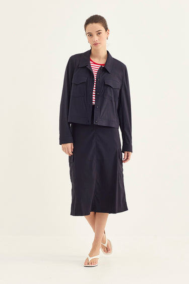 A model stands against a plain light background wearing a dark navy blue cropped jacket and matching midi skirt over a red and white striped top by Verge, paired with beige flat sandals.