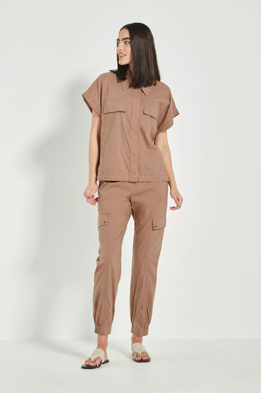 A woman modeling a matching two-piece outfit consisting of a short-sleeve button-up shirt and cargo pants by Verge in a warm taupe-brown color, set against a plain light gray studio background.