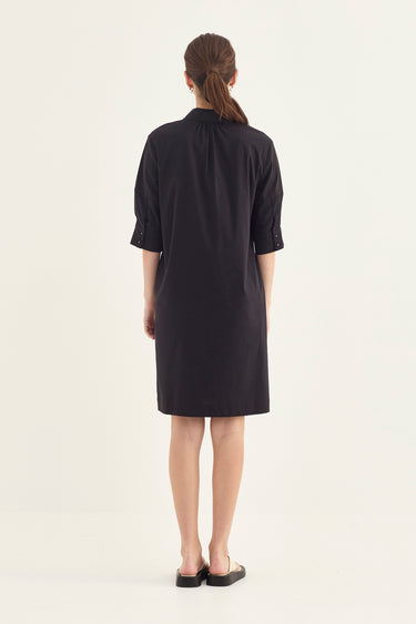 A woman viewed from the back wearing a black short-sleeve shift dress by Verge with rolled cuffs and black flat sandals, standing against a plain off-white studio background.