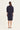 A woman viewed from the back wearing a dark navy blue shirt dress by Vergewith three-quarter sleeves and rolled cuffs, standing against a plain off-white studio background.
