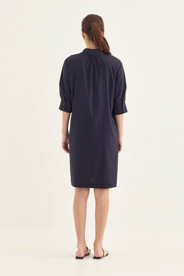 A woman viewed from the back wearing a dark navy blue shirt dress by Vergewith three-quarter sleeves and rolled cuffs, standing against a plain off-white studio background.