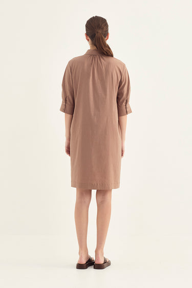 A woman viewed from behind wearing a short, loose-fitting brown dress by Verge with rolled sleeves and dark brown sandals, standing against a plain off-white studio background.