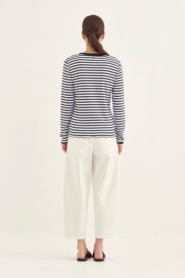A woman viewed from the back wearing a navy and white horizontally striped long-sleeve top, white cropped pants by Verge, and black sandals, against a plain light background.