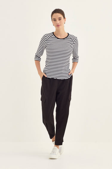 A woman modeling a black and white horizontally striped long-sleeve top with black cargo pants by Verge and white sneakers, standing against a plain off-white studio background.