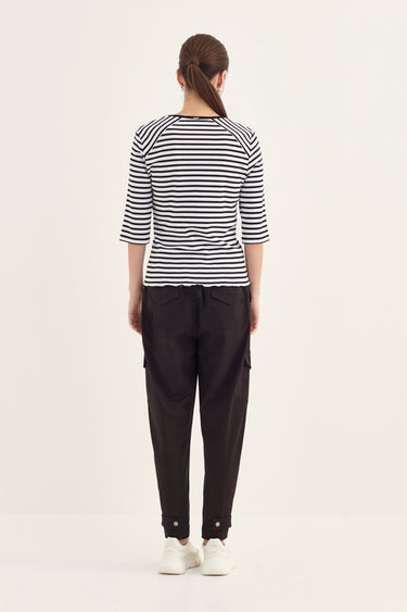 A woman viewed from the back wearing a black and white horizontally striped short-sleeve top, black pants by Verge, and white sneakers, standing against a plain light background.