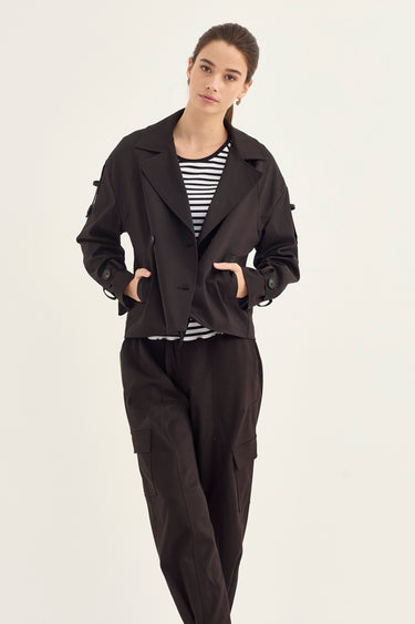 A woman modeling a black cropped utility jacket with side buttons, worn over a black and white striped t-shirt and black cargo pants by Verge, against a plain light background.