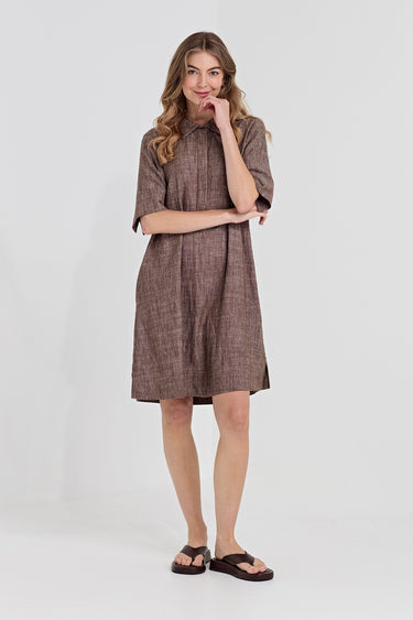 A woman modeling a short-sleeve brown linen dress by Verge with a textured weave, paired with dark brown slide sandals, against a plain light gray studio background.