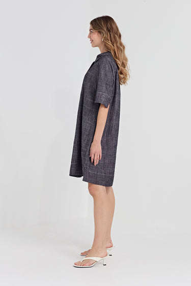 A woman modeling a short-sleeve, button-front shirt dress by Verge  in a dark gray textured fabric, shown from a side profile against a light gray studio background.