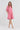 A woman modeling a pink short-sleeve linen dress with a high collar and side pockets by Verge, paired with white thong sandals, against a light gray studio background.