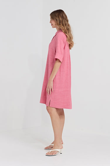 A woman wearing a pink short-sleeve linen dress by Verge with a relaxed, oversized fit and side slits, standing in profile against a clean white studio background.