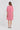 A woman viewed from the back wearing a pink short-sleeve textured dress by Verge with a collar and front pleats, paired with white heeled sandals, against a light gray studio background.