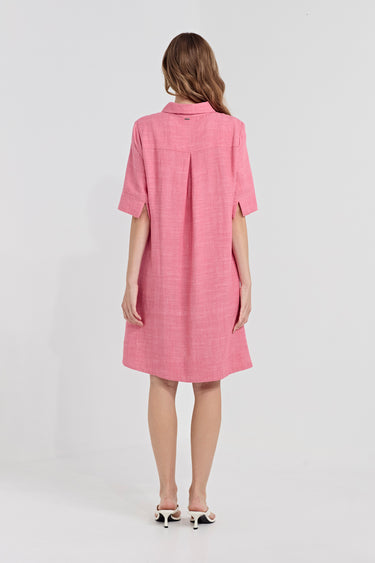A woman viewed from the back wearing a pink short-sleeve textured dress by Verge with a collar and front pleats, paired with white heeled sandals, against a light gray studio background.