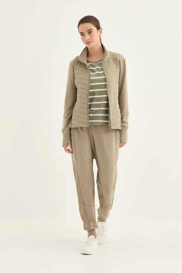 A woman modeling a light khaki outdoor tracksuit consisting of a zip-up jacket, a striped long-sleeve top, and matching jogger pants by Verge, paired with white sneakers against a plain white studio background.
