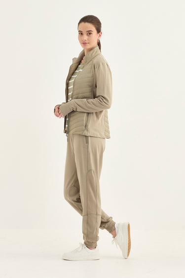 A woman modeling a matching beige athletic tracksuit consisting of a zip-up jacket and jogger pants by Verge, styled with a striped shirt and white sneakers against a plain white studio background.