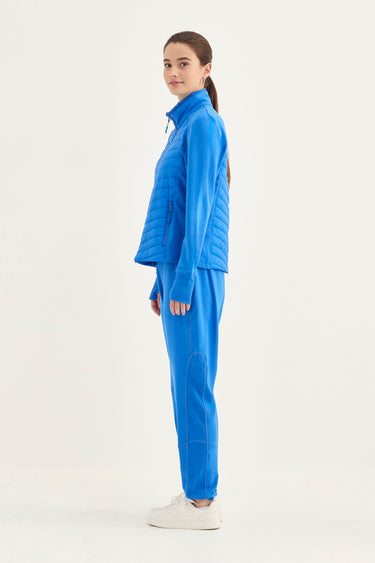 A woman modeling a bright blue matching tracksuit consisting of a quilted zip-up jacket and matching sweatpants by Verge, paired with white sneakers, against a plain white studio background.