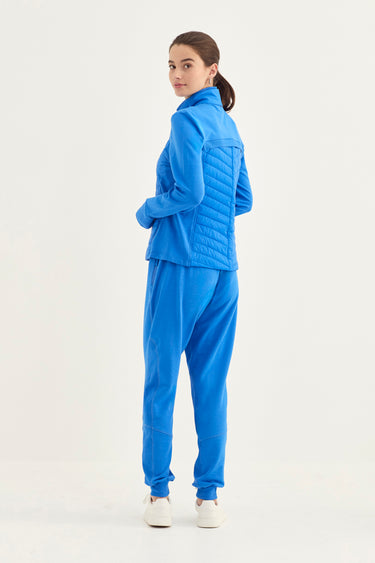 A woman modeling a bright blue matching set consisting of a quilted jacket and sweatpants by Verge, shown from a rear three-quarter angle against a plain white studio background.
