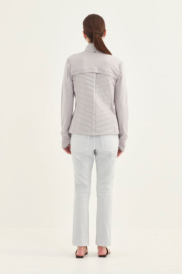 A woman viewed from the back wearing a light gray quilted long-sleeve jacket and white pants by Verge, against a plain white studio background.