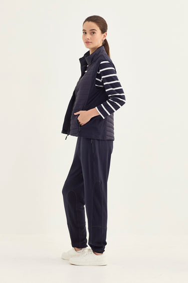 A woman modeling a navy blue puffer vest with navy and white striped sleeves, paired with matching navy pants by Verge and white sneakers, against a plain white studio background.