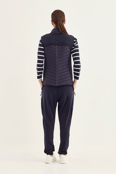 A model shown from the back wearing a navy blue quilted puffer vest over a navy and white striped long-sleeve shirt, paired with navy pants by Verge and white sneakers, against a plain light background.