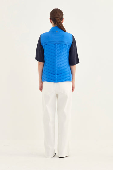 A model shown from the back wearing a bright blue quilted puffer vest over a black short-sleeve top and white wide-leg trousers by Verge, against a plain white studio background.