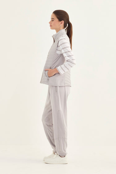 A woman stands in profile wearing a light gray sleeveless zip-front vest over a white and gray striped long-sleeve top, paired with loose light gray pants by Verge, and white sneakers, against a plain white studio background.