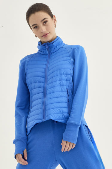 A woman modeling a bright blue cropped quilted zip-up jacket with a high stand-up collar, paired with matching blue sweatpants by Verge, against a plain light background.