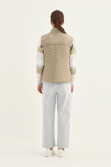 A model shown from the back wearing a beige quilted vest over a white and beige striped long-sleeve shirt, paired with light gray trousers and white sneakers by Verge, against a plain white studio background.