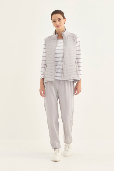 A woman modeling a light gray puffer vest over a gray and white striped long-sleeve top, paired with light gray jogger pants by Verge and white sneakers, against a plain white studio background.