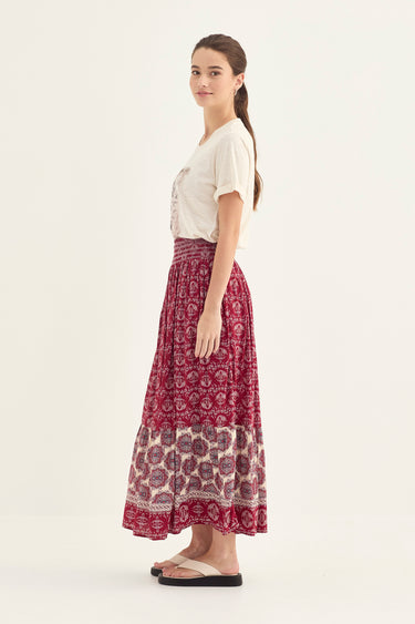 A woman stands in profile wearing a white short-sleeve top and a long, high-waisted maxi skirt by Verge with a burgundy and cream geometric pattern. She is wearing beige sandals against a plain, light off-white studio background.