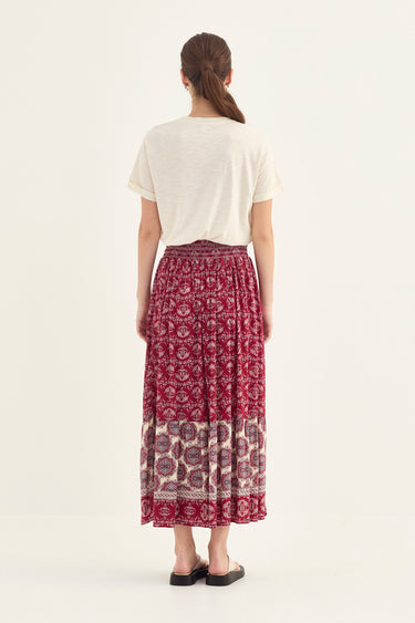 A woman shown from the back wearing a cream short-sleeve t-shirt and a long, pleated midi skirt  by Verge with a red and cream floral block print and a patterned hem, paired with black slide sandals, against a plain off-white studio background.
