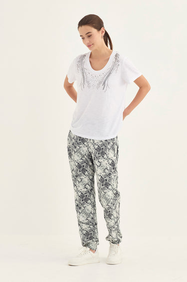 A woman modeling a white short-sleeve t-shirt with silver rhinestone embellishments on the chest, paired with black and white snakeskin print pants by Verge and white sneakers, against a plain white studio background.