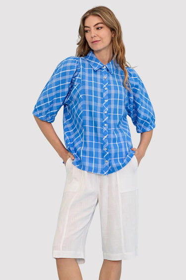 A woman modeling a blue and white plaid short-sleeve button-up shirt with puff sleeves, paired with white shorts by Verge, against a solid light blue background.