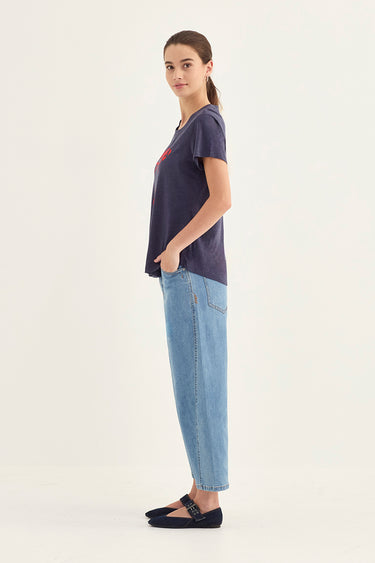 A woman stands in profile wearing a dark navy short-sleeve t-shirt with a small red graphic on the chest, light blue jeans by Verge, and dark navy flats, against a plain off-white studio background.