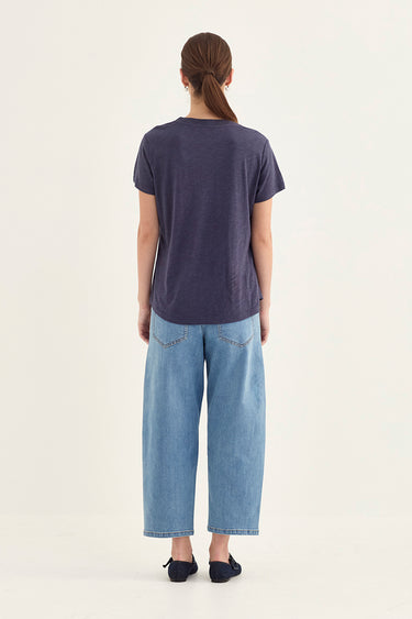 A woman viewed from behind wearing a dark slate blue short-sleeve t-shirt, light-wash wide-leg denim jeans by Verge, and black flats, set against a plain off-white studio background.