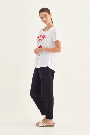 A woman modeling a white short-sleeve t-shirt with a red graphic print on the front, paired with black pants by Verge and white slide sandals, against a plain off-white studio background.