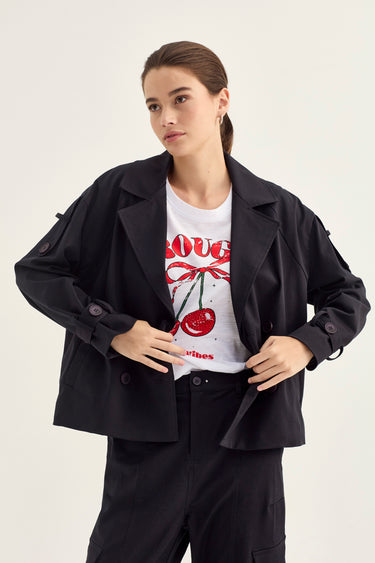 A woman modeling a black cropped blazer over a white graphic T-shirt with red cherry artwork and text, paired with black pants by Verge, against a plain light background.